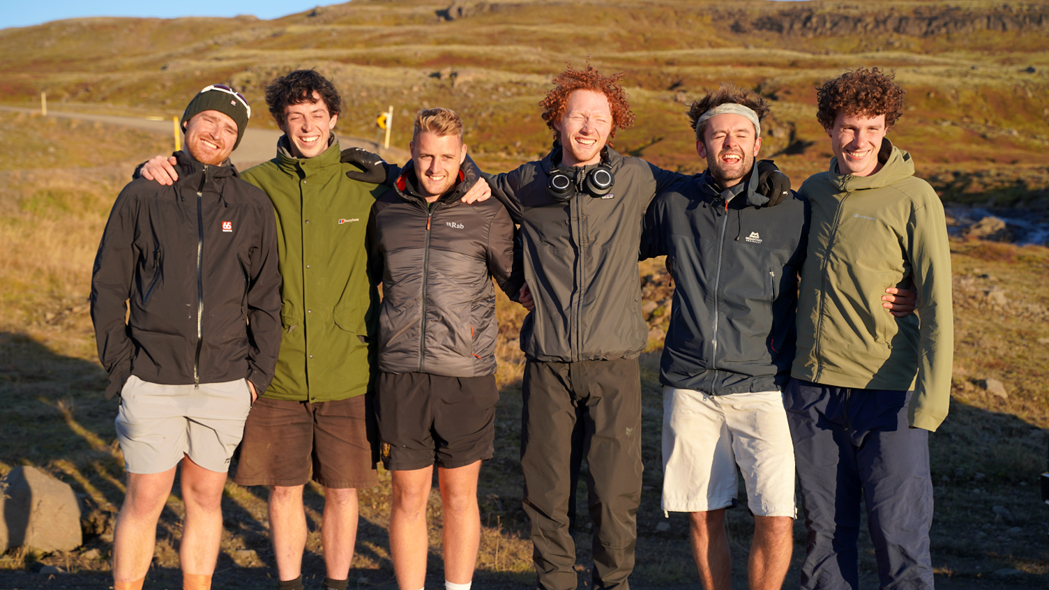 Six young men wearing waterproof coats and either activity trousers or shorts stand with their arms round each other smiling at the camera. In the background is a rugged-looking landscape of grass and exposed rock.