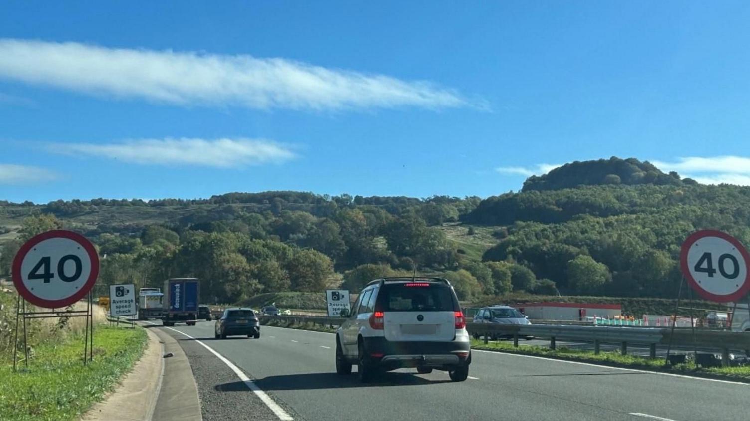A stream of traffic passes through a section of dual carriageway with signs saying that the road is limited to 40 miles per hour. Further back there are more signs letting drivers know that there are speed cameras in place. There is a white car in the foreground, wit ha black car further down the road and two lorries in front of that.