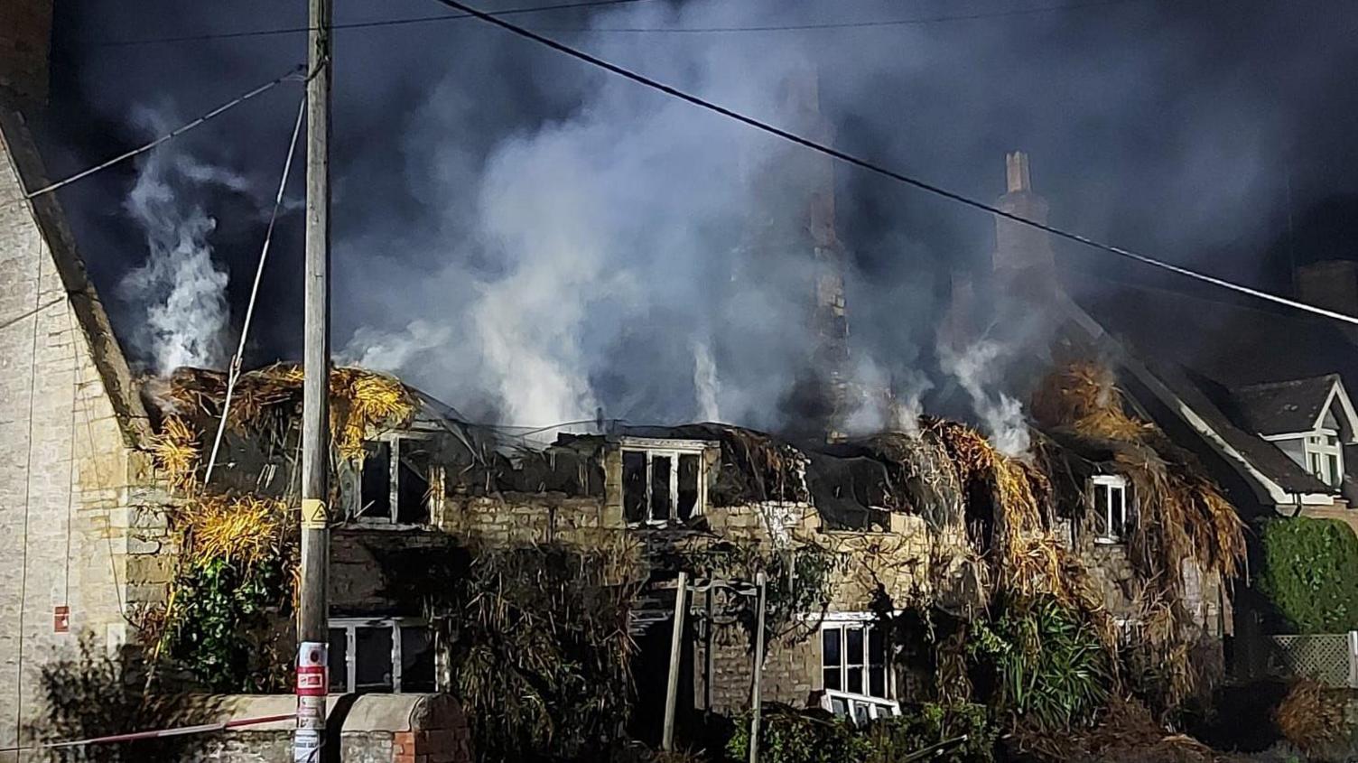 A burnt out home, that had a thatched roof, showing fallen thatch, and most of the the whole building destroyed. A large number of white wooden window frames, bushes outside the front and a small wall.