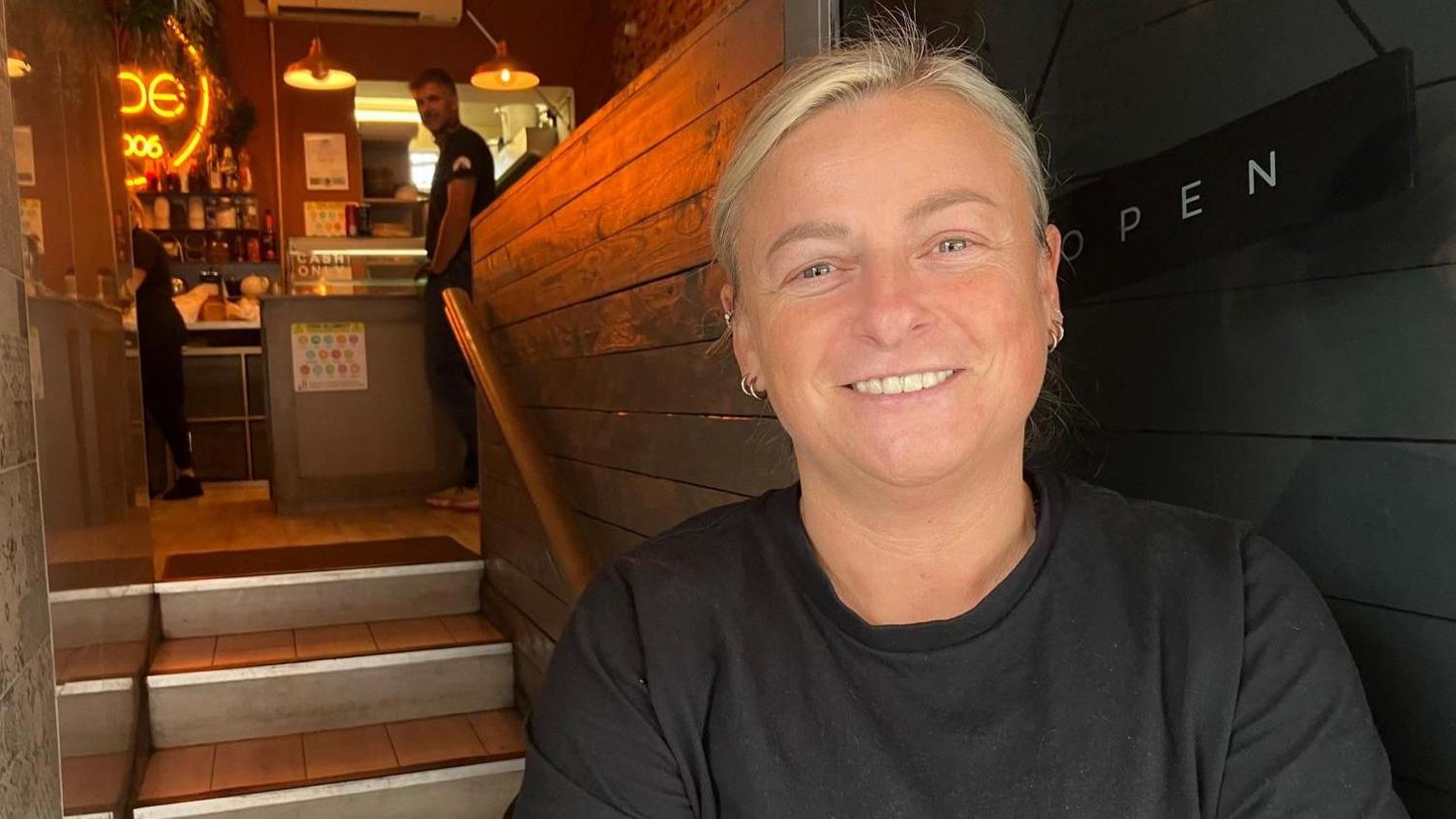 A long blonde haired woman has two ringed earrings in each ear. She stairs at the camera as she smiles in the doorway of a small building. She leans against a clear glass door, which separates her from a black wood wall. Over her right shoulder are three wooden stairs that lead to a café. A man with short black hair stands side on at the top of the stairs. He wears a black t-shirt and smiles.