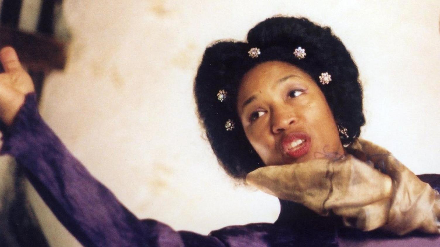 Mary-Ann Roberts yn perfformio ac yn gwisgo gemwaith yn ei gwallt, sgarff o gwmpas ei gwddf a thop porffor.