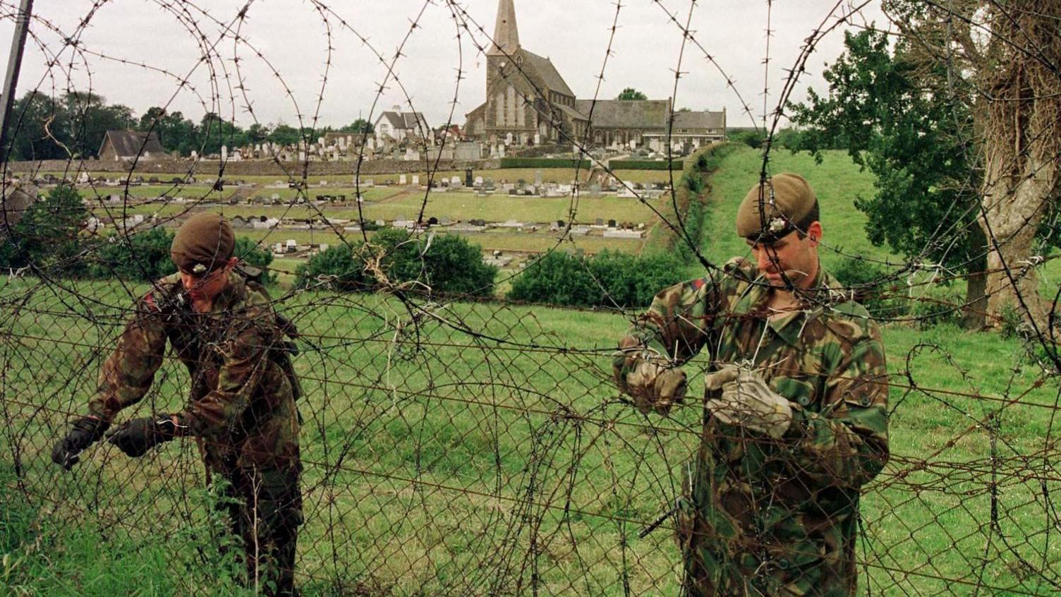Two British soldiers, dressed in camouflage, build a barbed wire fence in a field. 