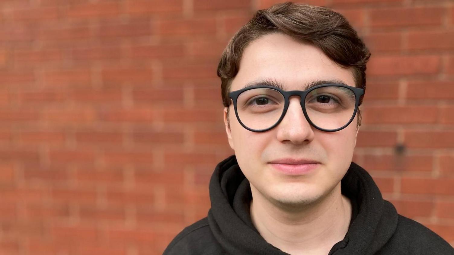 Mirzan looks directly into the camera in a close-cropped shot of his head and shoulders. He is wearing glasses and has medium-length wavy brown hair. He is wearing a dark coloured hoodie and stands in front of a blurred brick wall.