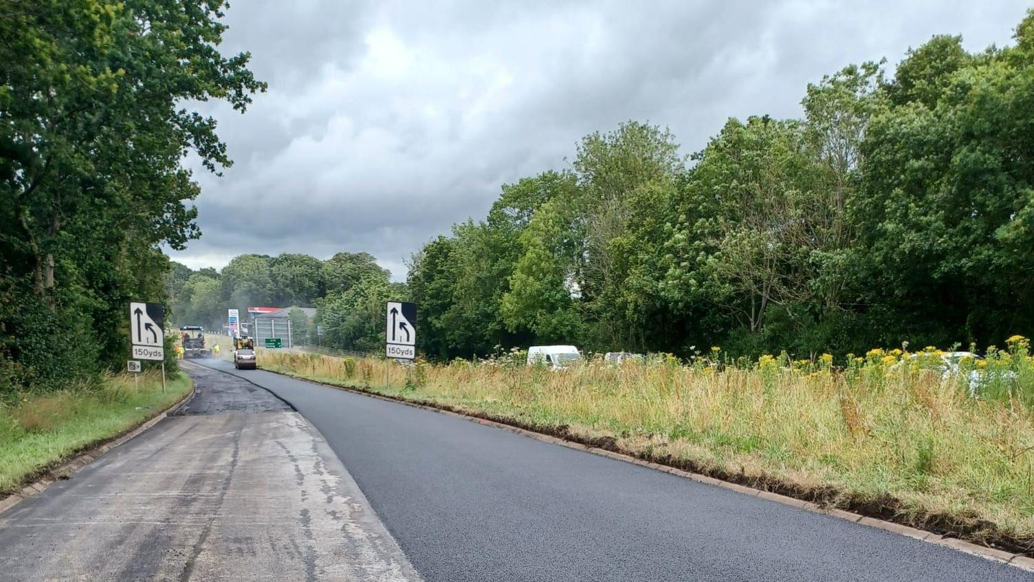 A steamroller is seen with a layer of tarmac being laid on top of the A24