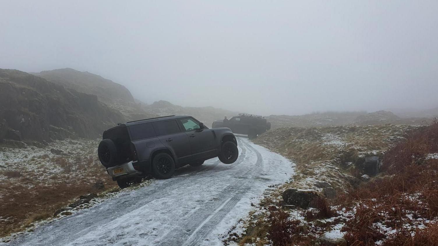 An icy single track road with two black cars run off it. The road is bordered by tall mountains.