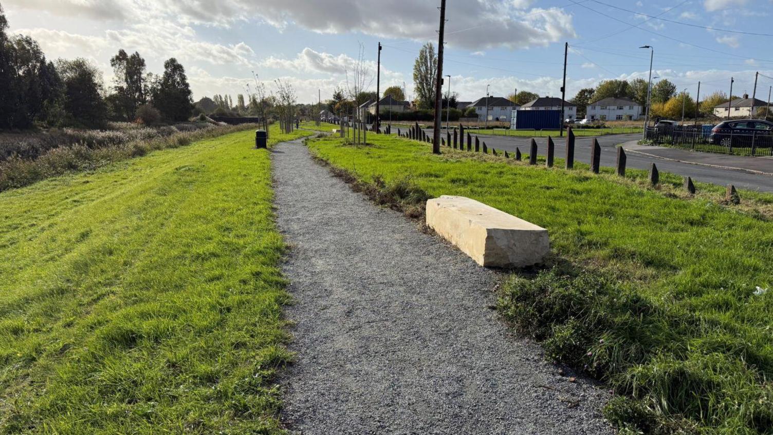 A gravel path surrounded by grass. There is a large stone seat next to the path. It is next to a road, and there are houses on the other side of the road.
