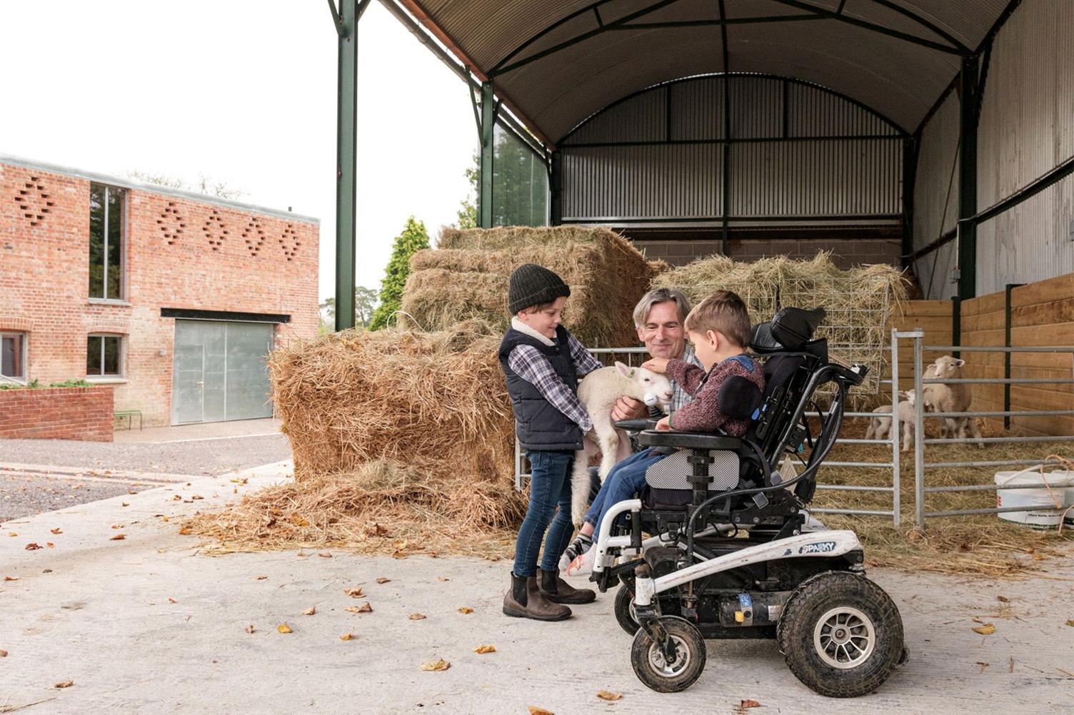 Inside a barn at Wraxall Yard, two boys - one in a wheelchair - are petting a sheep, which is being held by an older man