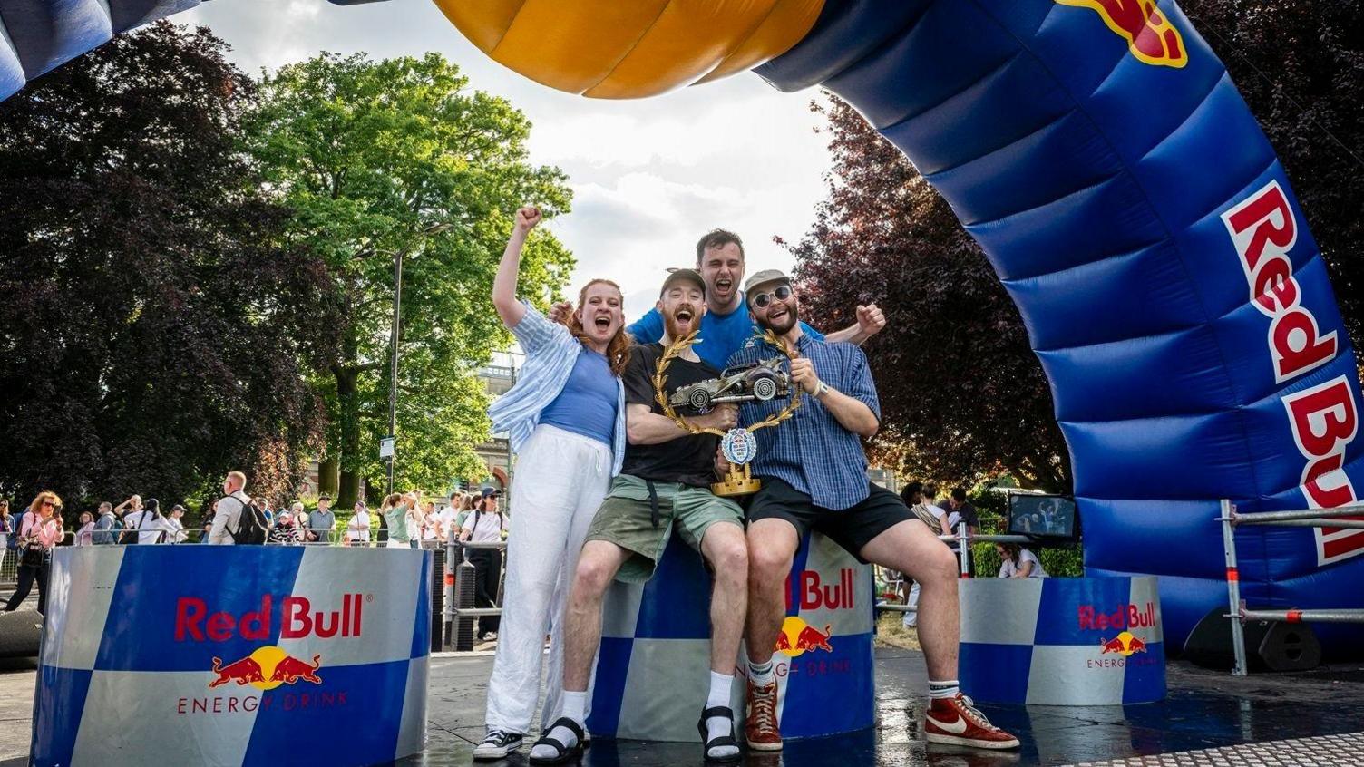 The Hurry Houdinis team, made up of three men and a woman, celebrate winning the race, holding up their trophy under an arch with Red Bull branding