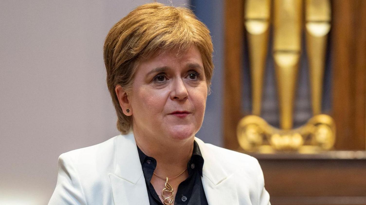 Nicola Sturgeon wearing a white jacket and black shirt holding her hands out in front of her as she talks