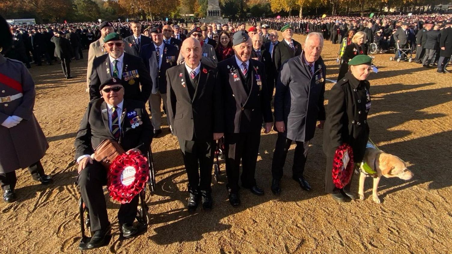 John Hardy and other veterans pictured at a Remembrance Sunday parade. He is in a wheelchair holding a poppy wreath. A lady on the other end of the line is walking with a guide dog and holding a poppy wreath. Four men are stood between them dressed in suits and ties.