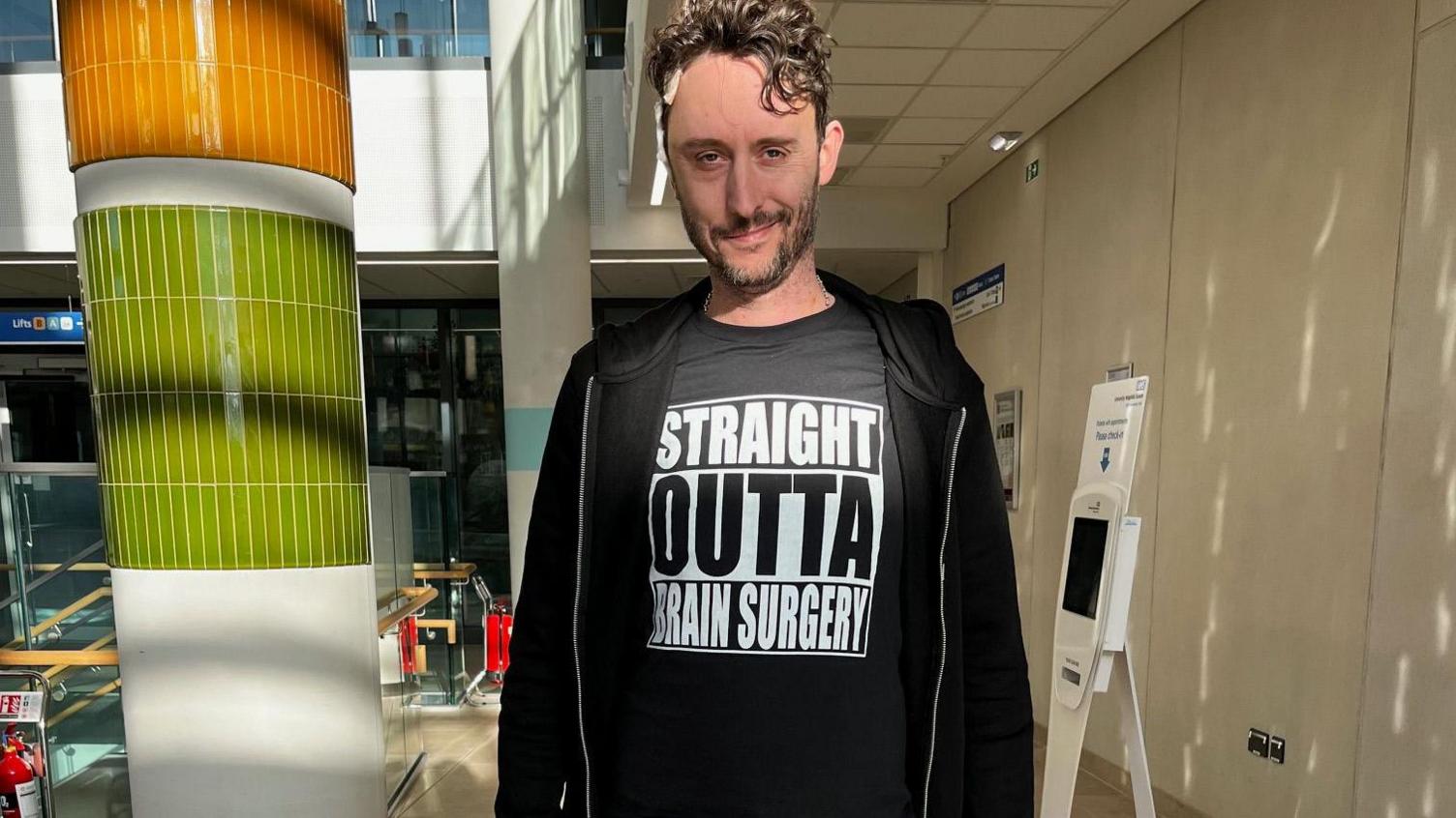 Matthew White in hospital, wearing a hoody, PJ bottoms and a t-shirt saying STRAIGHT OUTTA BRAIN SURGERY.