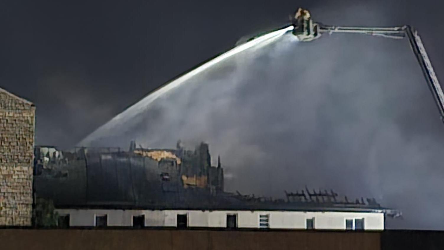 A firefighters can be seen on an aerial ladder platform working to extinguish the blaze. The charred blackened and badly damaged roof of the building can be seen.