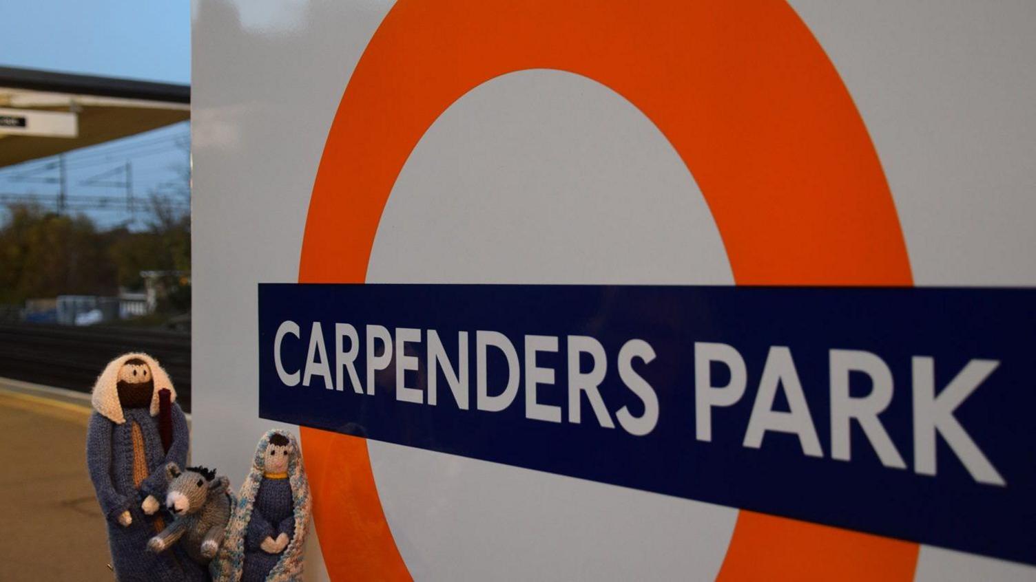 Miniature knitted figurines of Mary, Joseph and a donkey outside Carpenders Park Station sign.