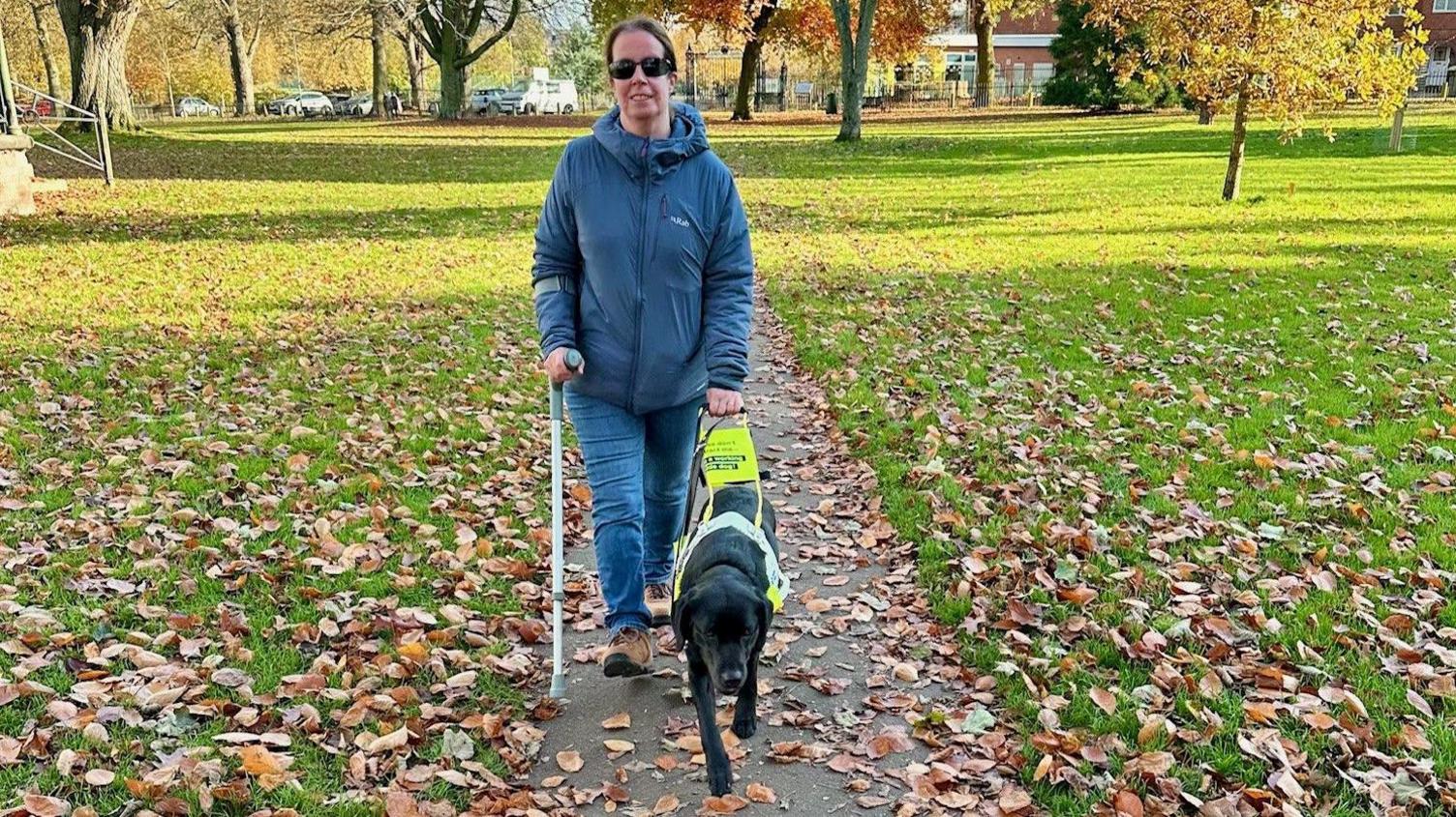 A woman and her guide dog walking on a path through a park. The woman is wearing sunglasses, wearing a coat and holding a crutch and the lead attached to her guide dog - a black Labrador. 