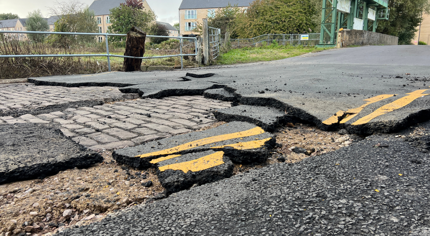 Images shows Mill Lane road and chunks of tarmac with yellow lines on them broken up with the cobbles underneath exposed.
