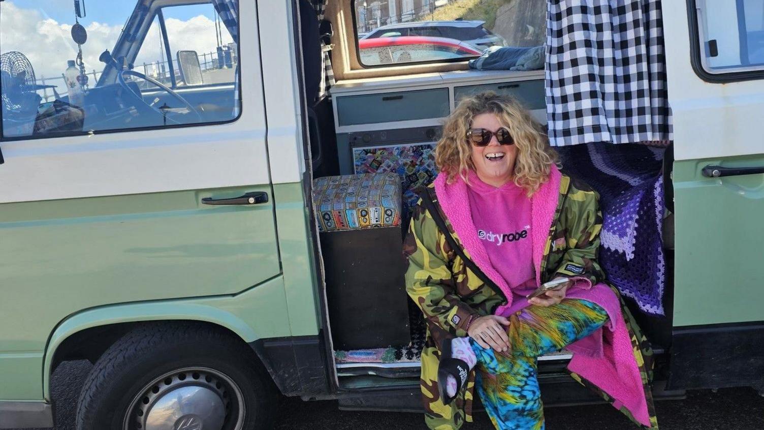 Susan Universe sitting in the van. She is wearing a green camouflage coat and a pink jumper. She has blonde hair and is wearing sunglasses. A black and white chequered curtain and knitted blanket can be seen inside the van.