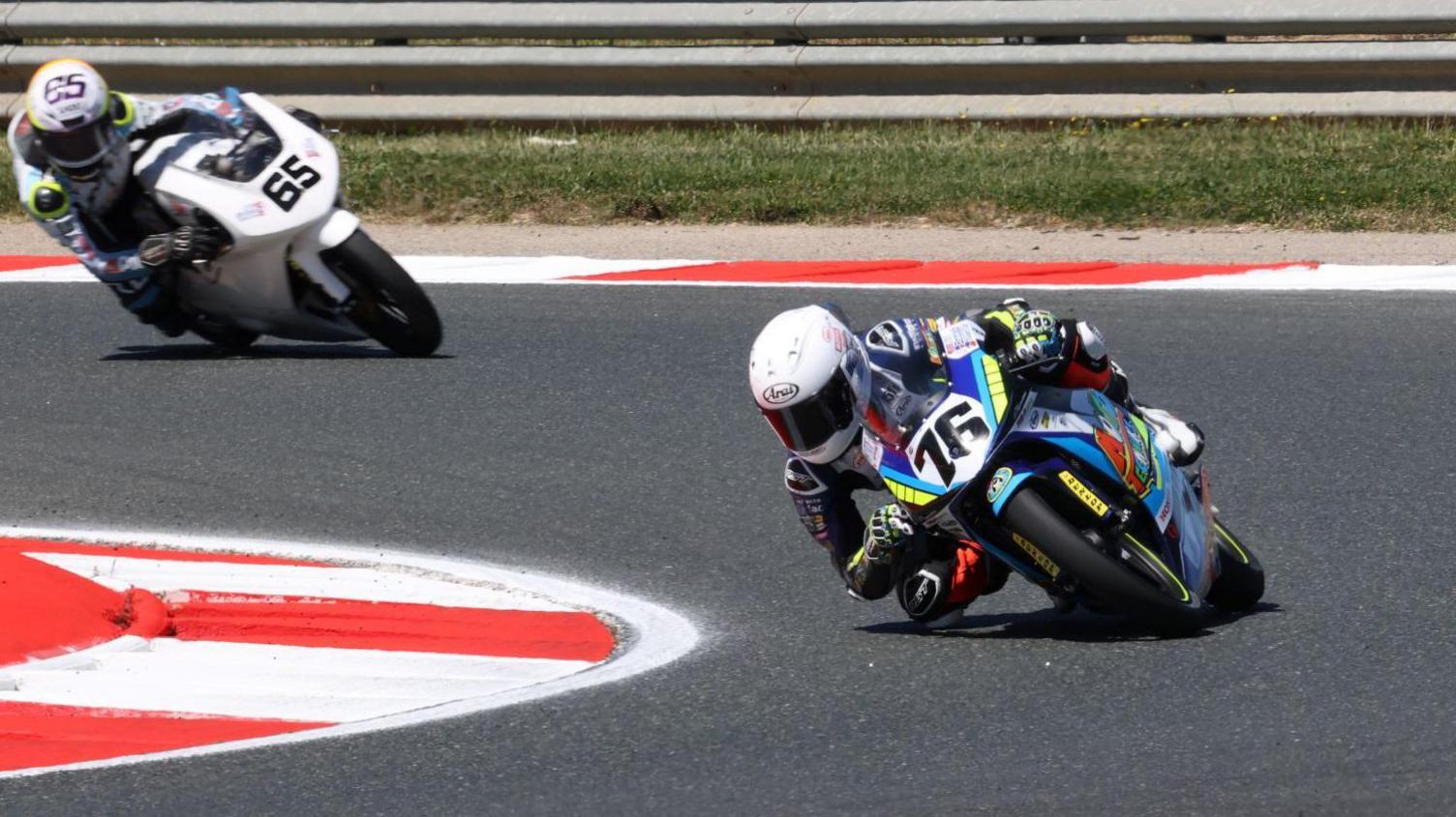 Charlie Huntingford racing his bike, which has the number 76 on it, on a track with another bike close behind him. He is wearing a race suit and white helmet. 