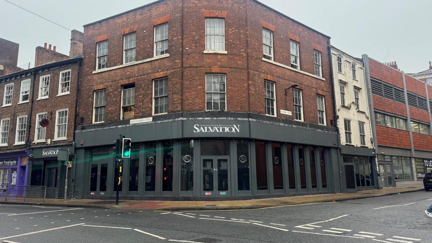 A three-story building on the corner of a asphalt road junction. The ground floor is grey painted window frames, the next two floors are red brickwork. White letters above a ground floor door entrance read Club Salvation.