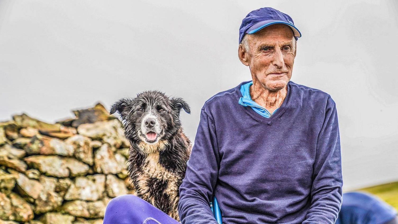 Joss Naylor sitting on a Lake District fell, accompanied by a dog