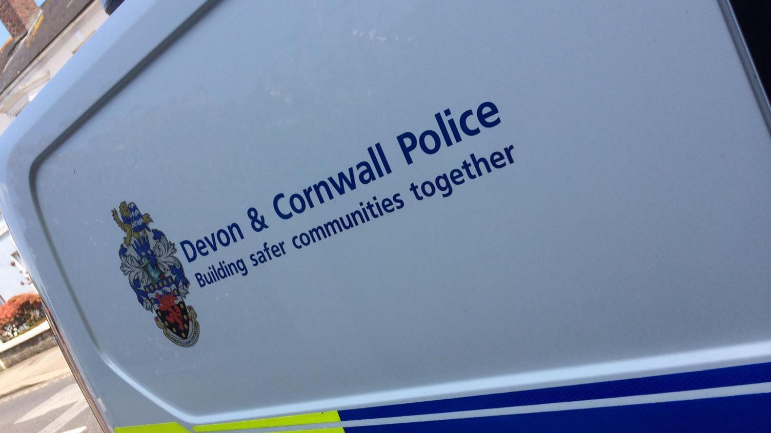 The side of a blue and green police vehicle. It has the Devon and Cornwall Police logo with writing next to it. There is a slogan that reads - 'Building safer communities together'