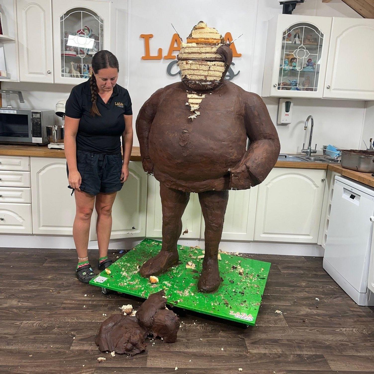 A woman with brown hair plaited over her shoulder is wearing a black shirt and shorts. She is looking at the floor. She is standing next to a large cake sculpture which is covered in chocolate ganache. The sculpture's face has fallen to the floor.