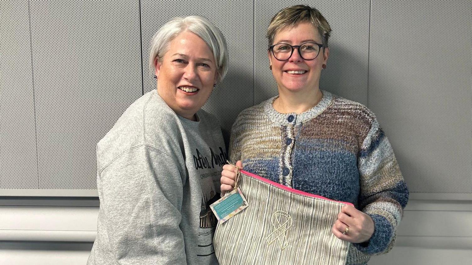 A woman with short grey hair and wearing a grey jumper, and a woman with short blonde hair, glasses and a multicoloured stripy cardigan. The woman wearing glasses is holding up a beige bag with a pink zip and a ribbon logo on the front. They are both smiling at the camera.