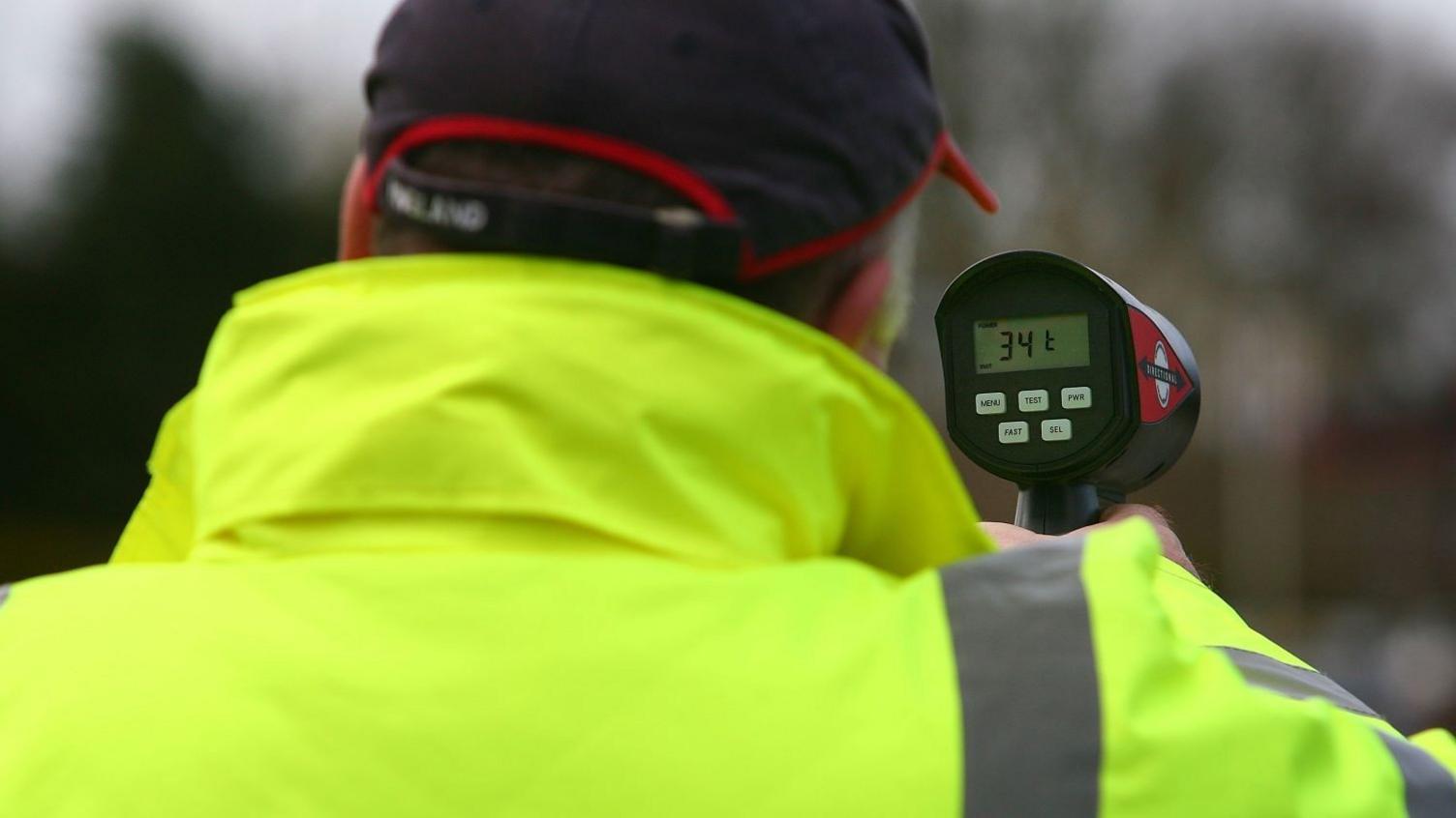 An over-the-shoulder photo of someone wearing a cap and high-vis jacket while holding a speed gun. The gun's display reads "34.1"