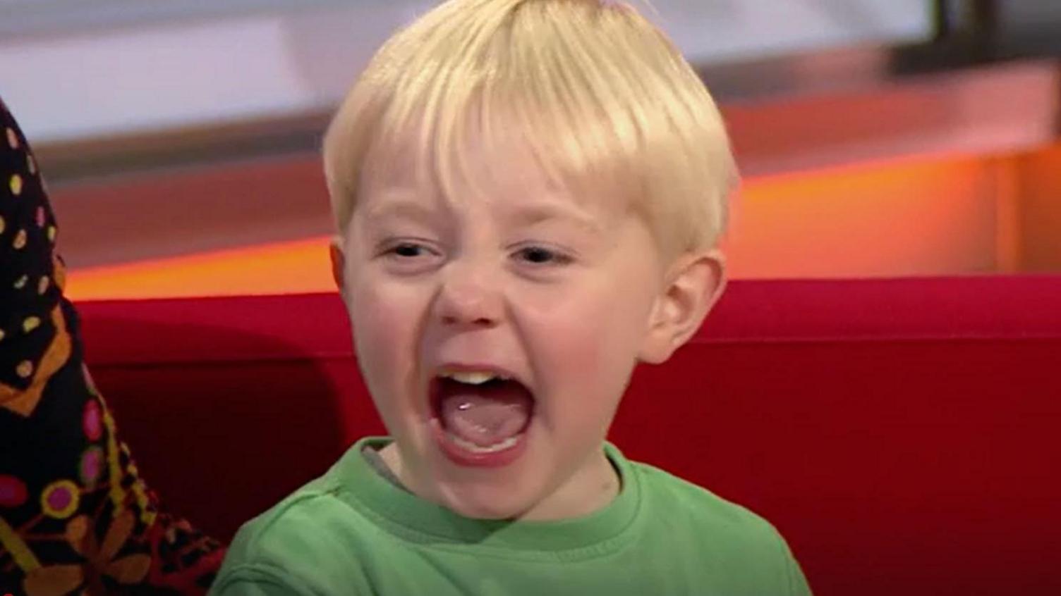 A young boy with blond hair and a green tshirt with his mouth wide open and a red background