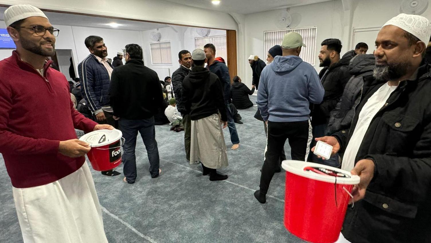 A group of men in a mosque, two men are holding red buckets for donations. Several men are behind with their back to the camera. 