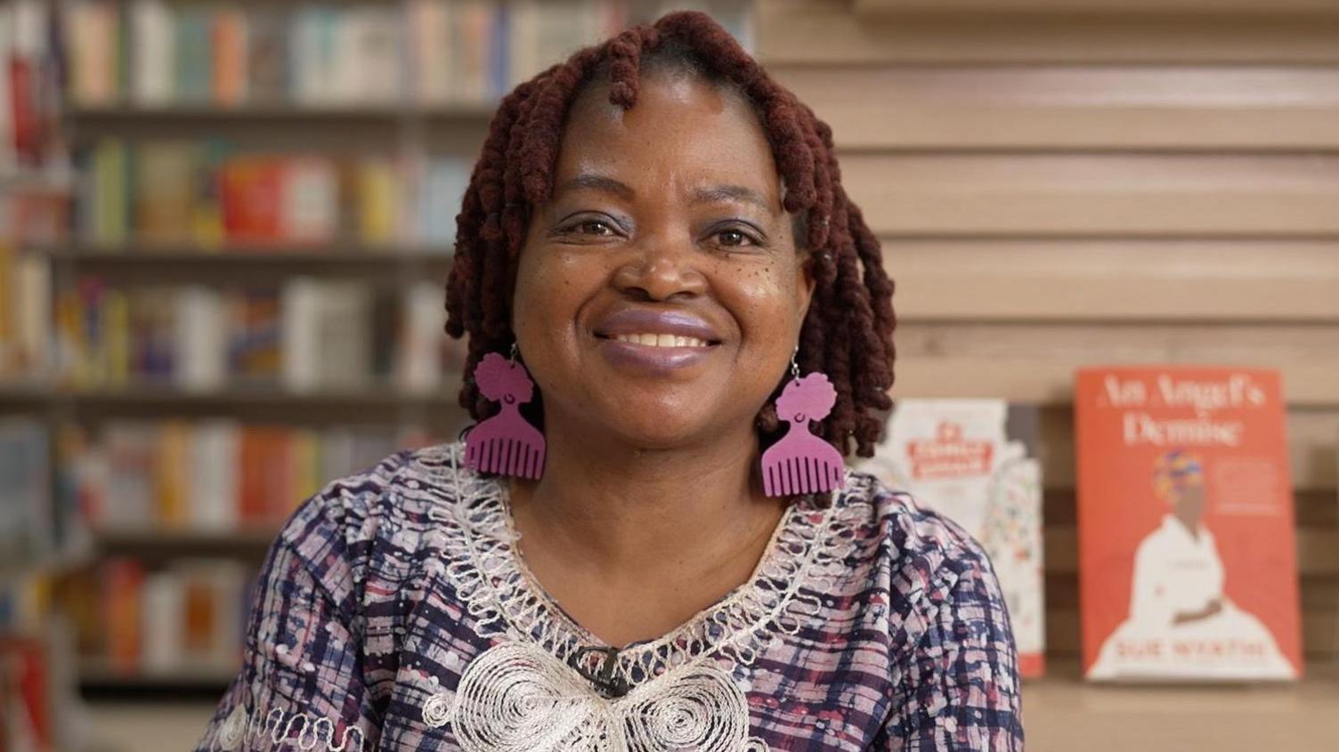 A woman with reddish-brown hair and pink comb-shaped earrings smiles at the camera, wearing a pink and blue checked top with white embroidered trim. She's in a bookshop, with blurred shelves of books on the left and a display featuring copies her books on the right.