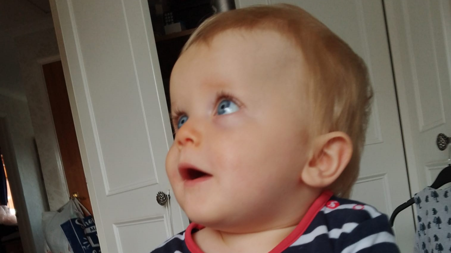 A one-year-old baby in a stripy top sits in front of some white cupboards looking up to the right of the room. 