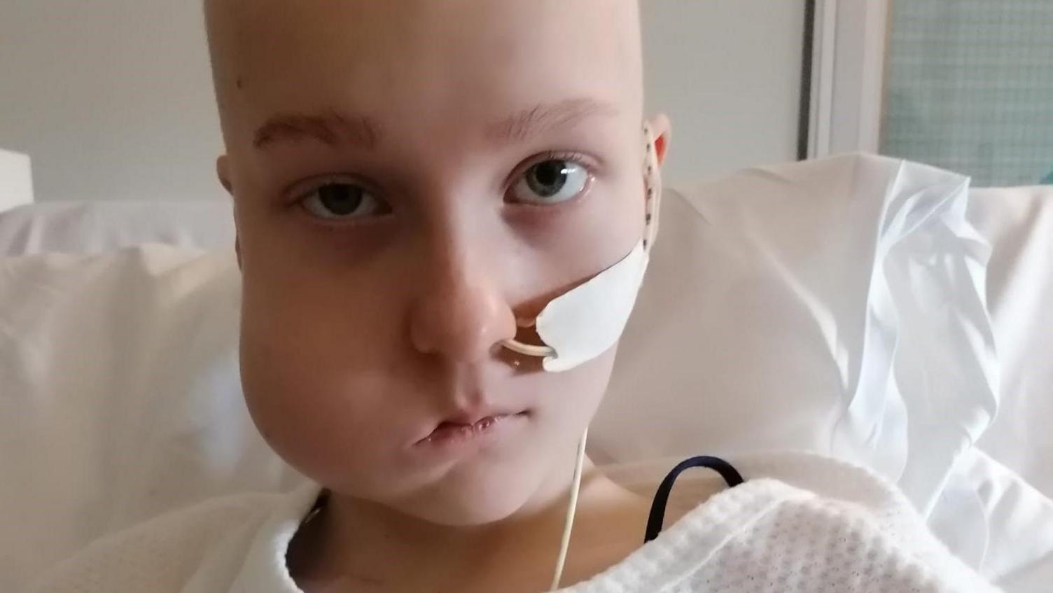 Gaby is on a hospital ward in bed. She has no hair and the tumour in her jaw muscle is visible. There is a tube running up her neck and into her nose which has been taped to her face. She is not smiling as she takes the photo of herself.  