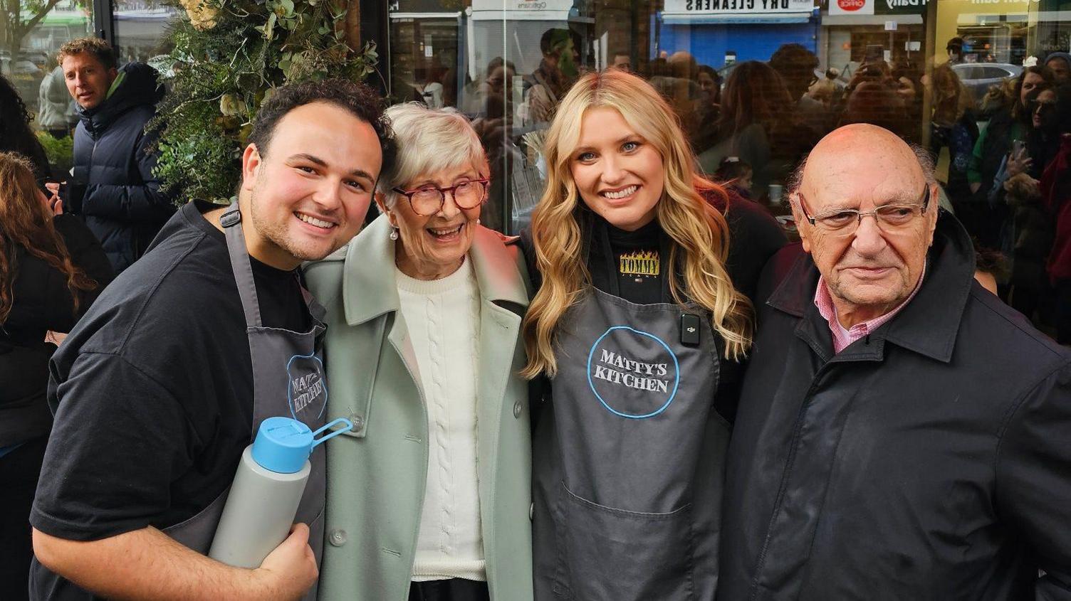 Matty and Ella wearing "Matty's kitchen" grey aprons in a group photo with June and Robert, a couple in their 90s