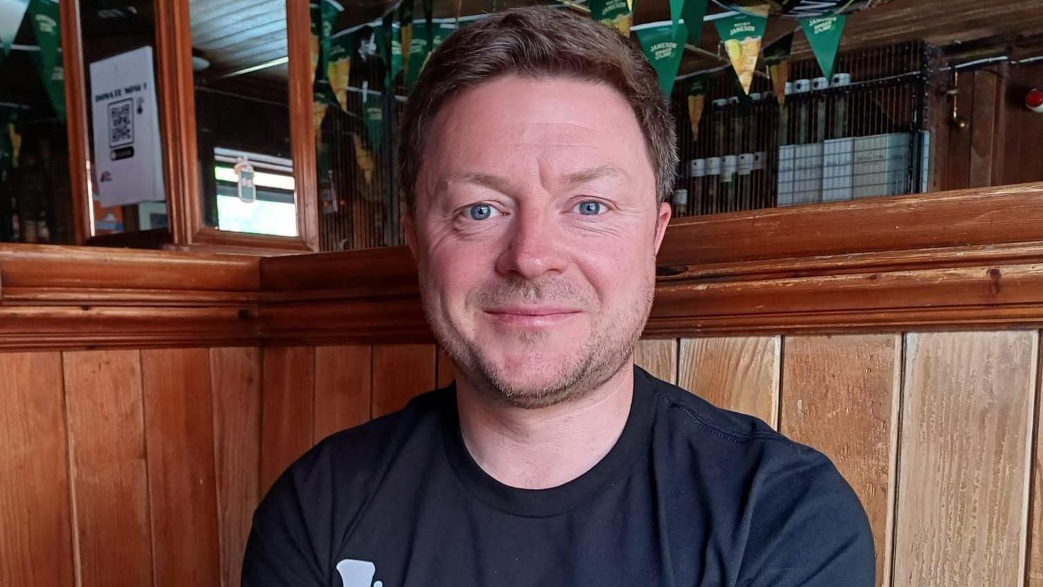 Donál is sitting in a bar. He's looking straight at the camera and wearing a black t-shirt