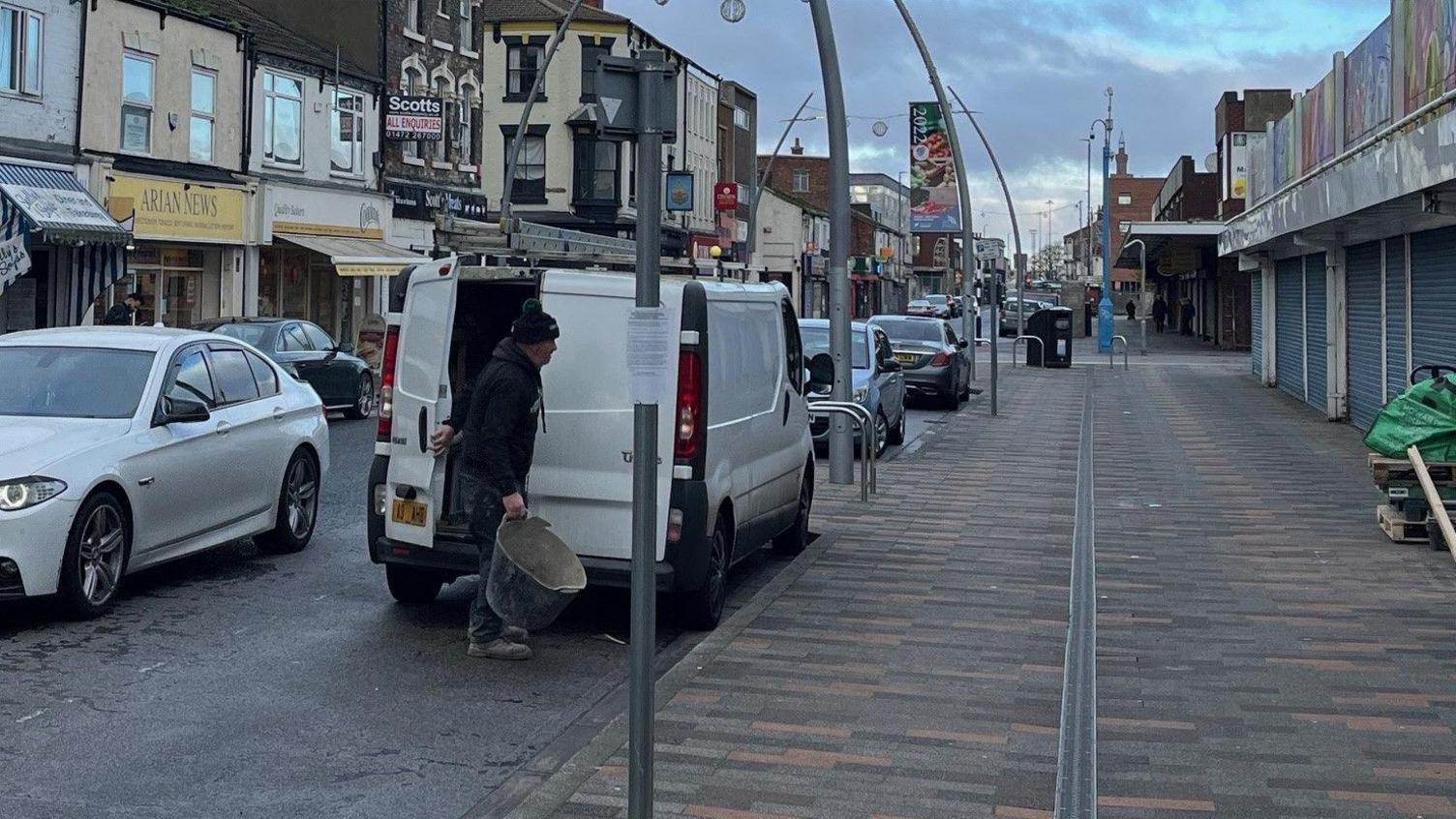 Dave Johnson carries a bucket from his white van parked in Freeman Street, Grimsby. Shops, some closed, are on both sides of the street. Dave is wearing dark clothes and a woollen hat