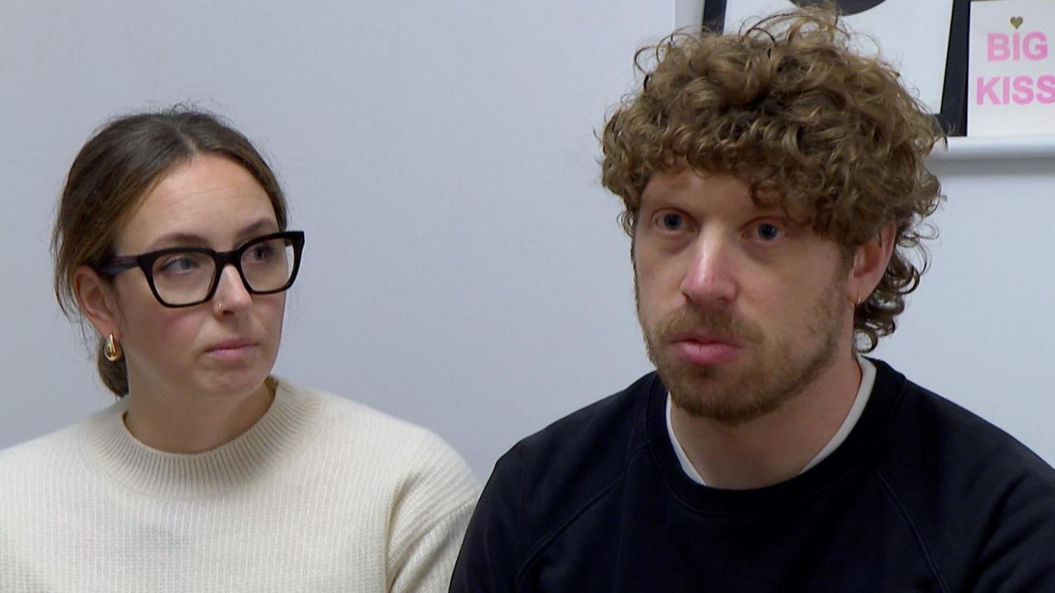 A man with curly brown hair and a black jumper on looking off camera next to a woman with a white jumper on and black glasses. 