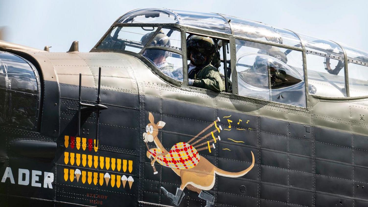 Three crew-members look out from the glass cockpit of a Lancaster bomber. The plane is painted partly in brown and green camouflage, and partly in black. A logo shows a cartoon kangaroo playing the bagpipes.