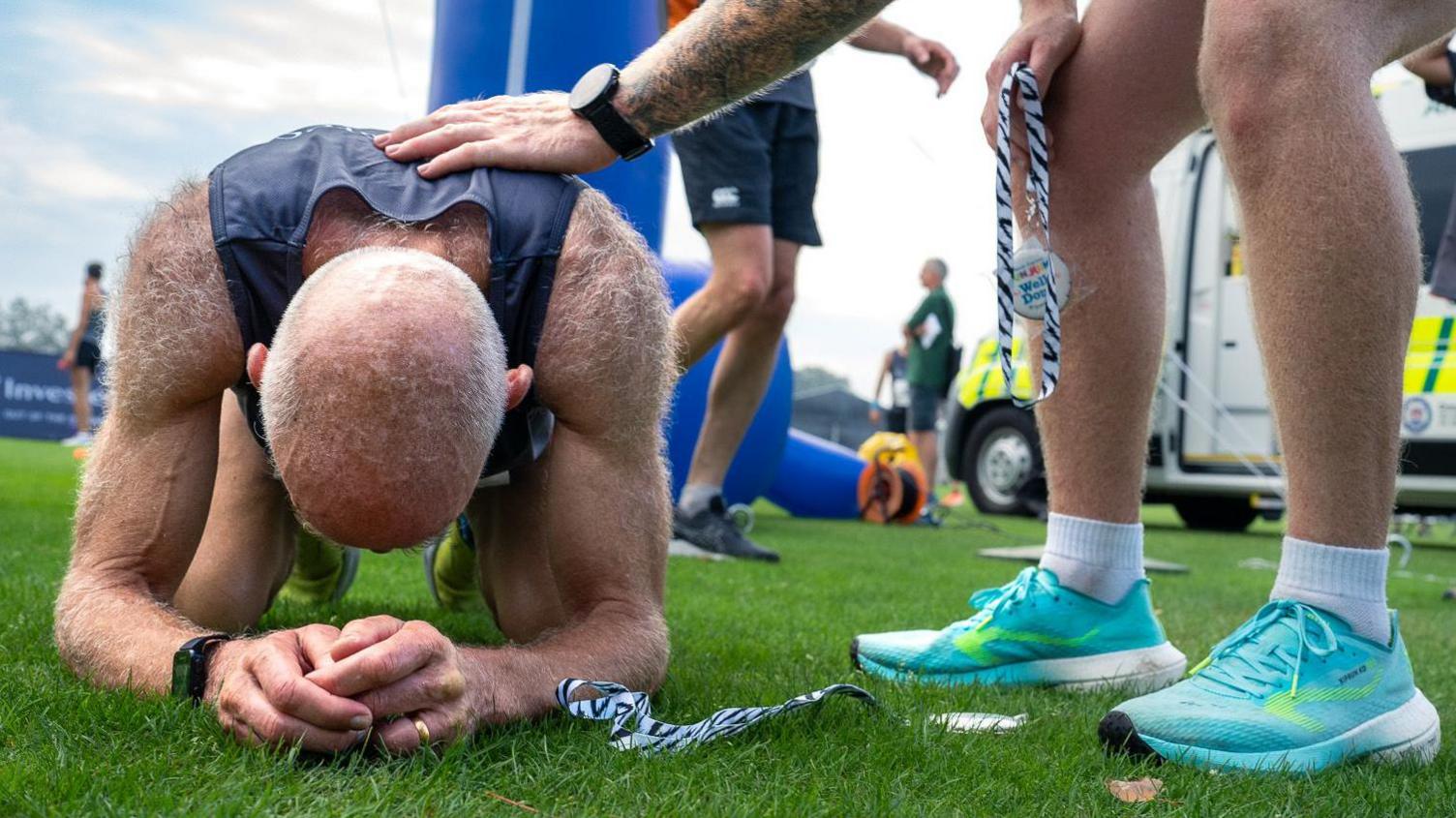 A male runner, muscles tensed, recovers on all fours on the grass at a previous 10K Challenge in aid of Mind Guernsey. His hands are clasped in front of him and the top of his head is facing the camera, while a comforting hand from another competitor rests on his shoulder.