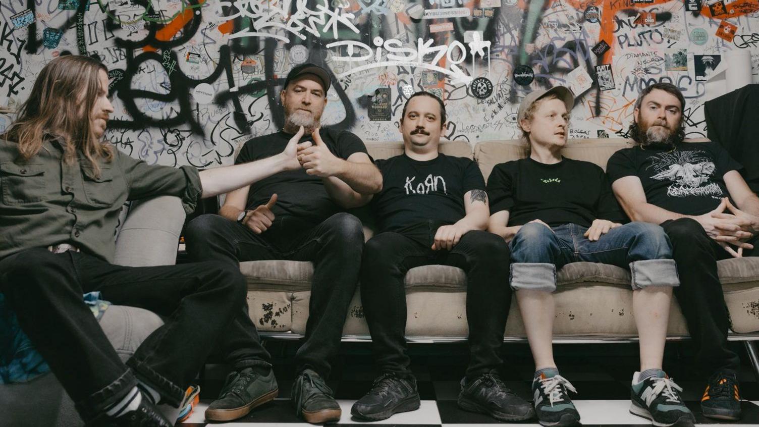 Pigs x7 which is made up of five men, all sitting on a sofa in front of a graffitied wall. They are all wearing dark colours and the two on the left are holding their thumbs together like they're in the middle of a thumb war. The man on the far left has long, brown hair and is looking to the side. Next to him, the man has a grey beard and is wearing a black cap. The man in the middle has dark hair which looks to be tied up and a dark moustache. He is wearing a black Korn band t-shirt. The man middle right has light hair and is wearing a grey cap and is looking to the right with his hands in his lap. He is wearing turned up jeans that are exposing the bottoms of his legs. The band member on the far right has chin-length dark hair and a greying beard and is looking into the camera with his hands clasped on his lap. 