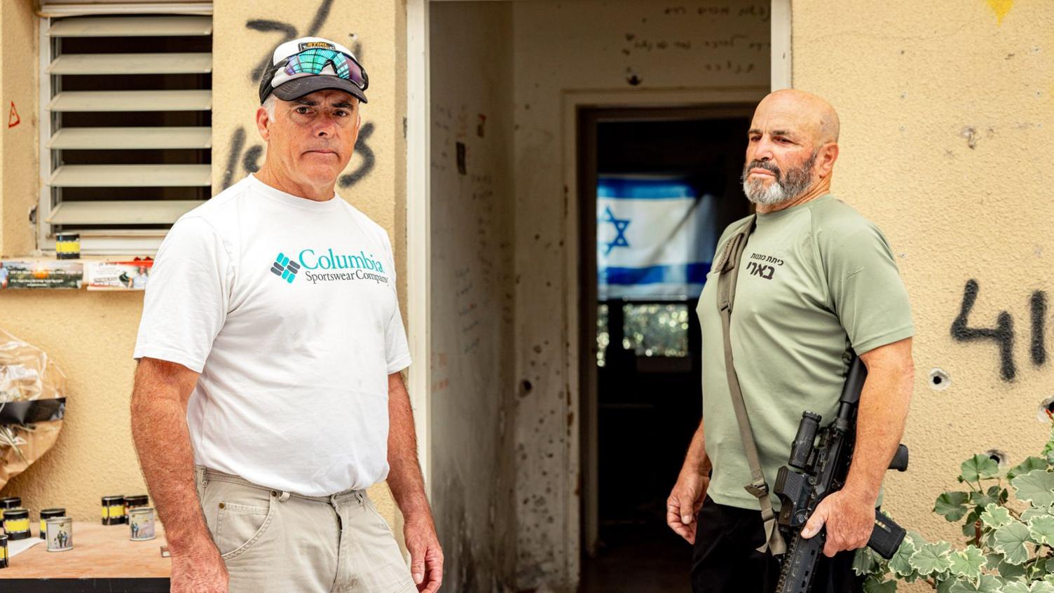 Two men. One aged 60 - the other 70. One is clean-shaven wearing a white T-shirt and cap. The other has a beard, is wearing a green T-shirt and is carrying a gun