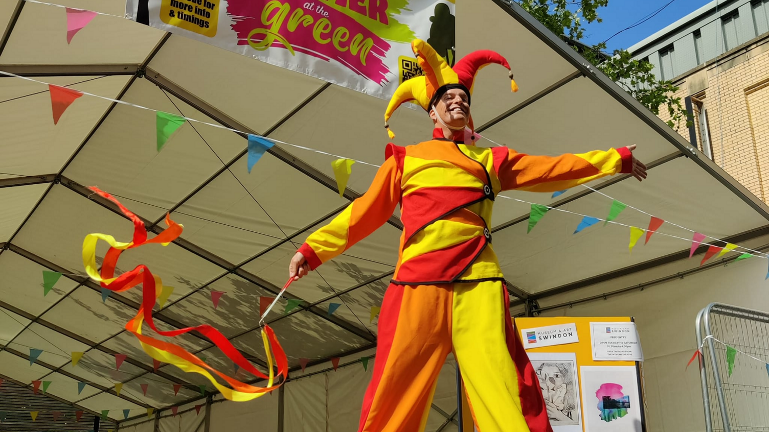 A man on stilts in a jester-type outfit coloured red, yellow and orange, holding a stick with ribbons. There is bunting and a white marquee behind him.