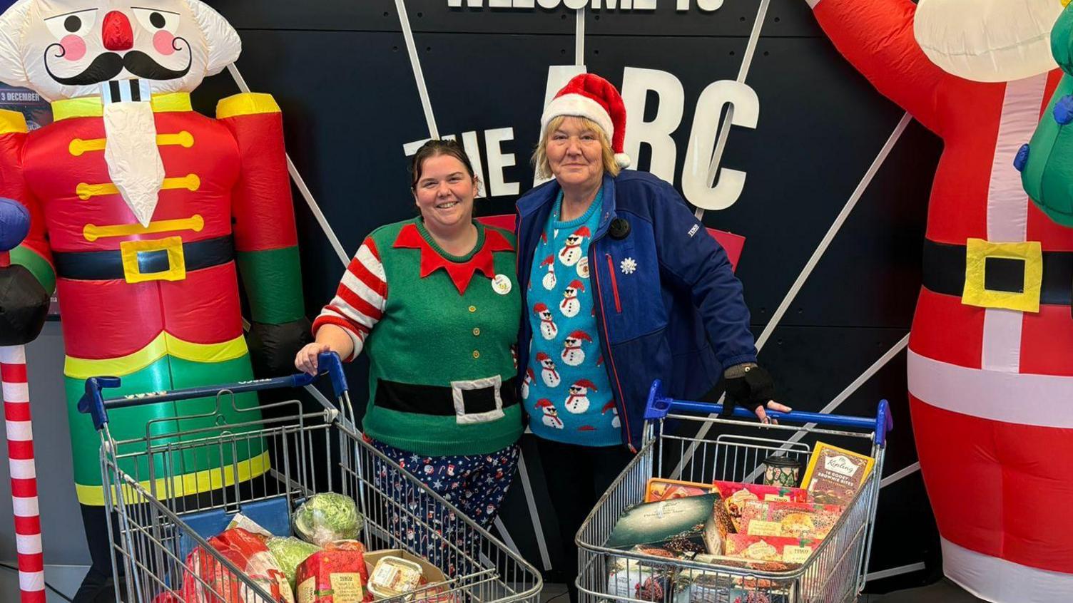 Two supermarket trollies filled with Christmas foods, including a turkey and mince pies. Two women in Christmas jumpers - one green and one blue with snowmen on them - hold the trollies. There are two inflatables - a Santa and a soldier - on the edge of the frame. 