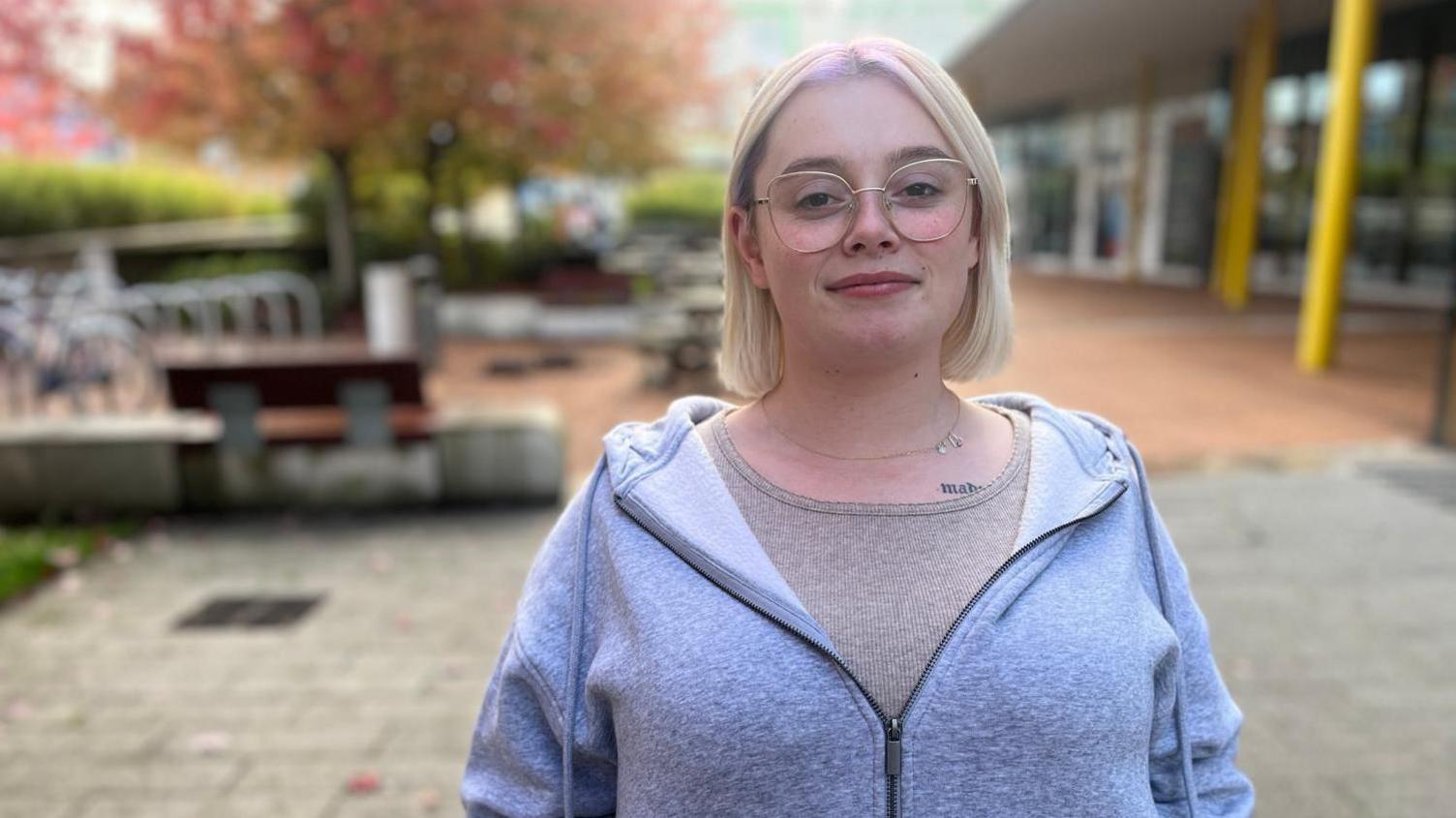 Katie is smiling into the camera stood outside a university building, wearing a grey zipped hoody over a beige top. She has shoulder length blonde hair and is wearing glasses.