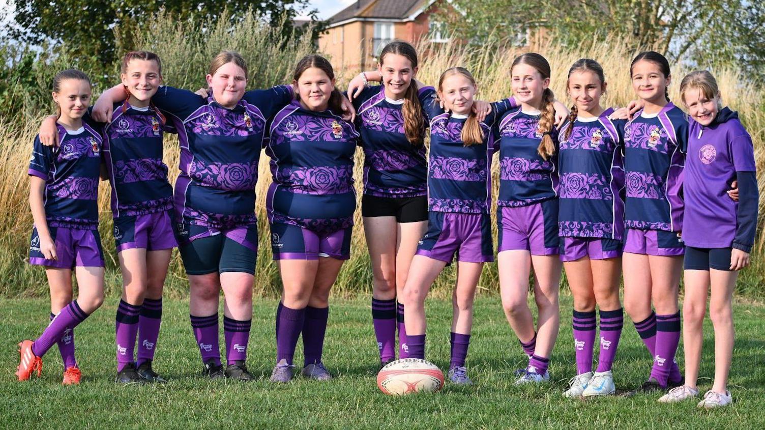 Ten girls, all under the age of 12, stand in a line on a grass pitch in their striped purple jerseys with their arms around each other's shoulders. They smile at the camera, with a rugby ball on the grass in the middle of them.