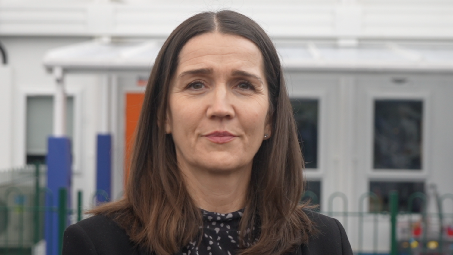 Academy trust chief Jo Coton in front of a white school building. She has long brown hair and is pouting while wearing a black blazer and navy and white top.