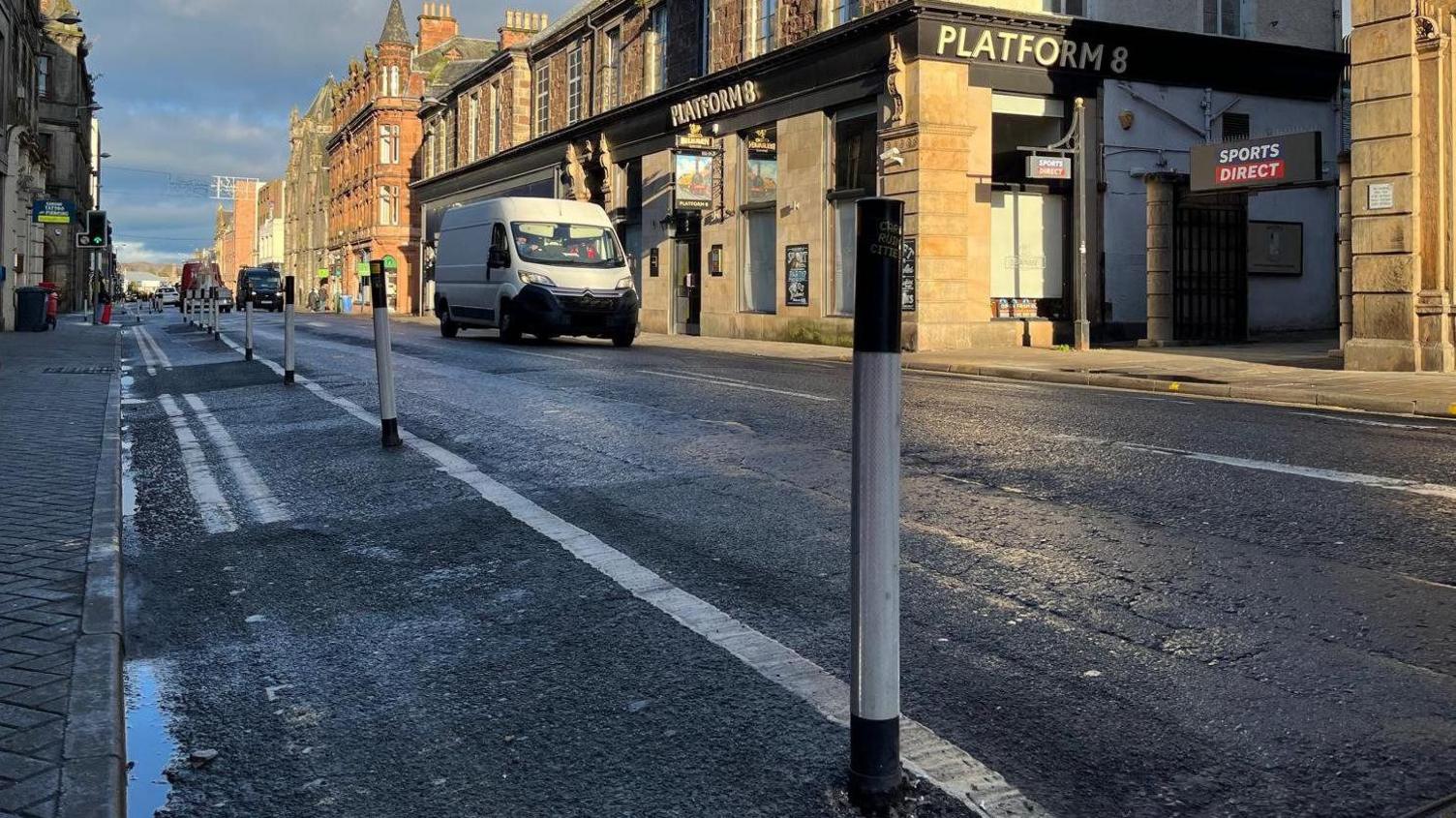 A low-level view of black and white bollards, running down the left hand side of Academy Sreet. In the background, a white van is parked outside a pub called Platform 8. 