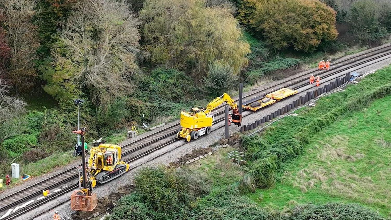 Yellow diggers are on a railway track where on one side a wall made of metal planks is being constructed. There are workers in hi-viz on the tracks as well and trees seen on the far side of the track