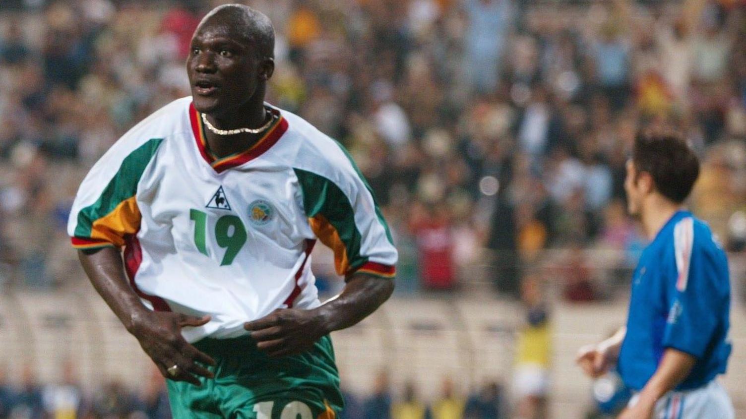 Senegal's Papa Bouba Diop, visible from waist up, is seen running away in celebration after scoring a goal against France in the opening match of the 2002 World Cup. Diop wears green shorts and a white shirt with green, yellow and red trim, which he looks like he is untucking at the waist. A green number 19 is positioned in the centre of the shirt while a big gold chain bounces around Diop's neck. In the background is a French defender in a blue shirt looking away from the camera and a big crowd blurred in the background