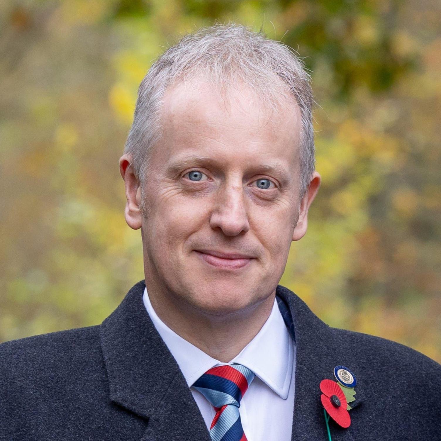 A man with short white hear wearing a dark coat with a poppy pinned to it, with a white shirt and red and blue tie.