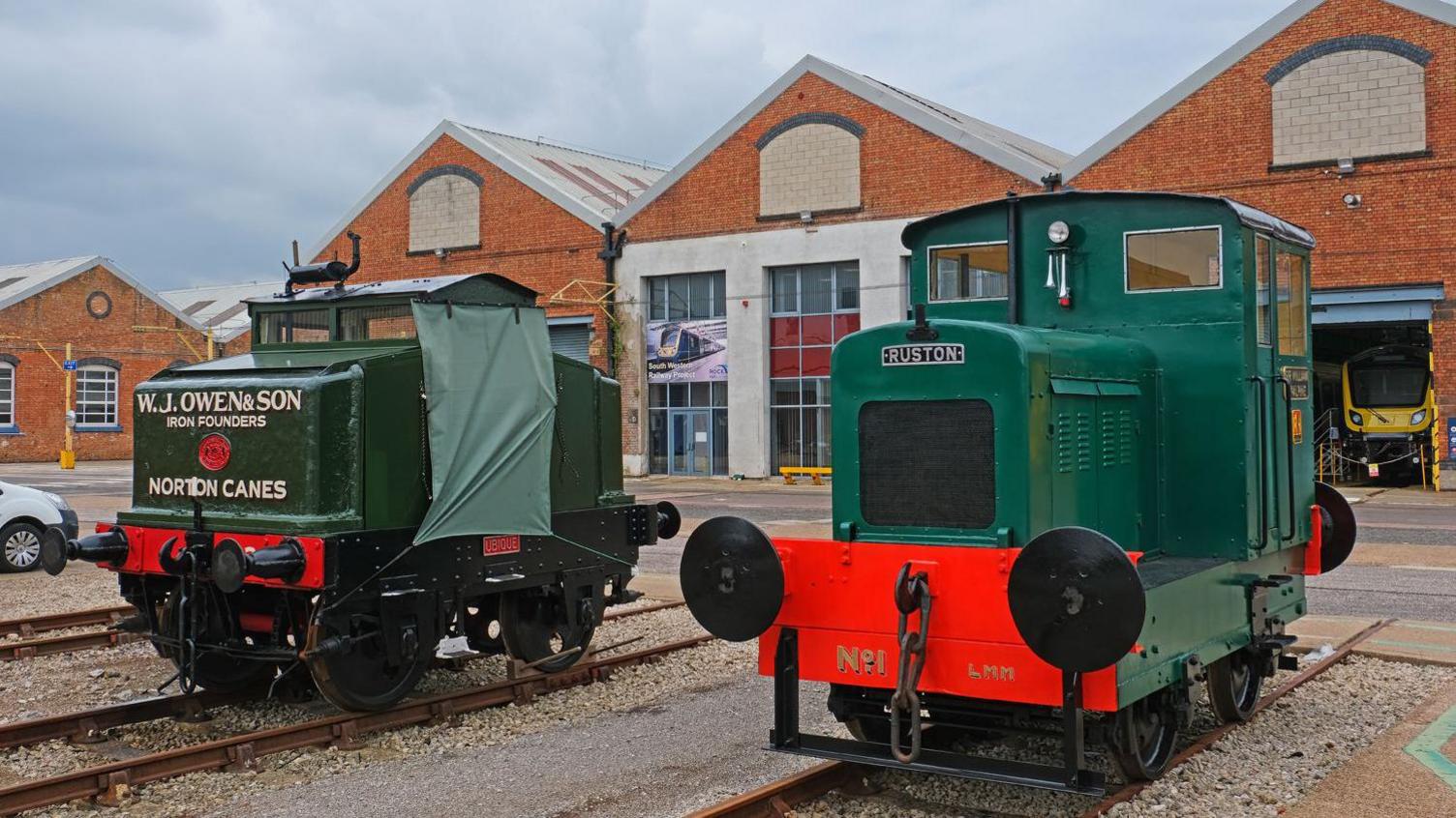 Two vintage rail vehicles are displayed outdoors, with red brick buildings in the background